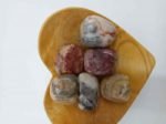 A heart-shaped wooden bowl with a variety of tumbled CRAZY LACE AGATE stones inside.