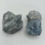 Due pezzi di Calcite Blu Grezza Naturale su una superficie bianca.